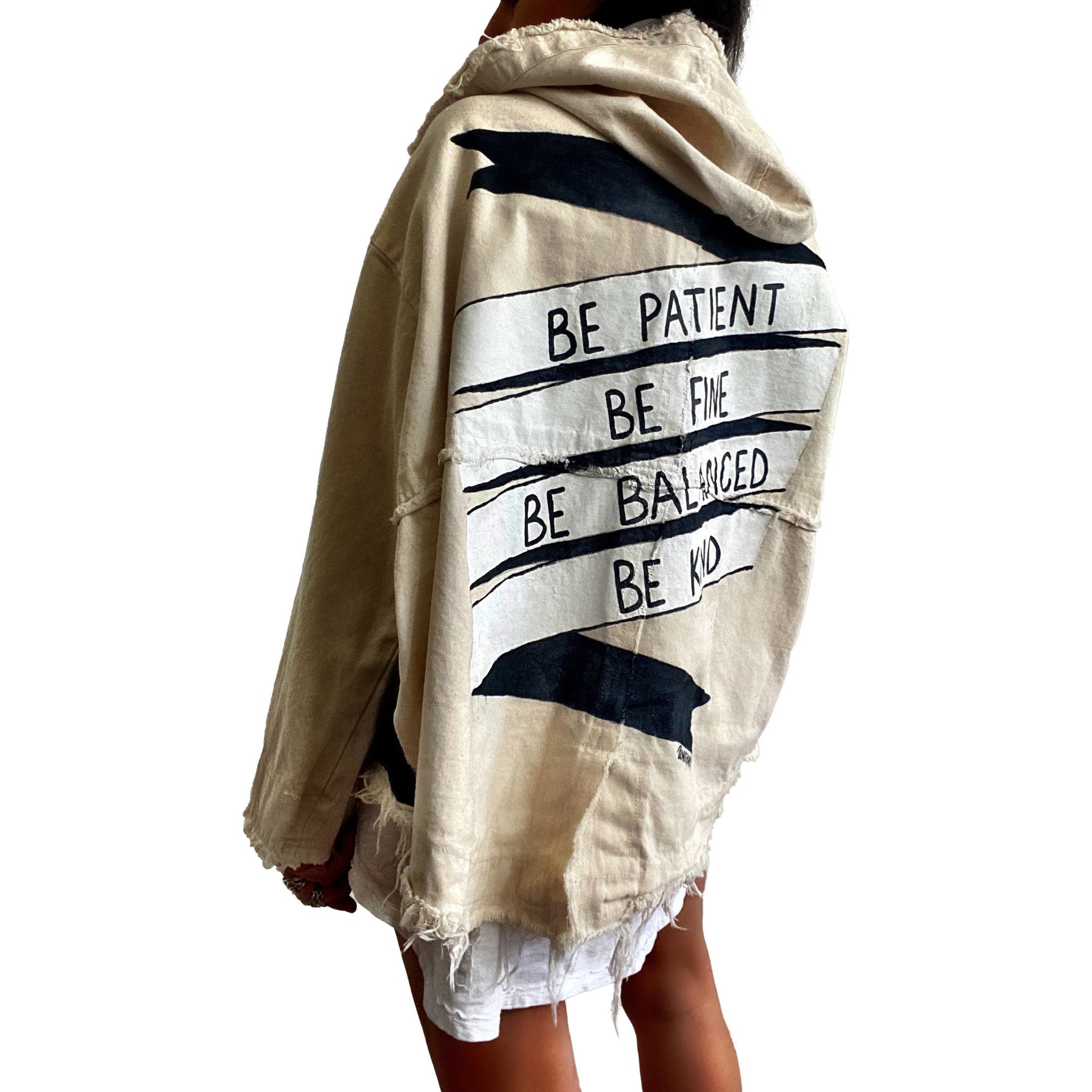 Beige hooded denim jacket. BE PATIENT, BE FINE, BE BALANCED, BE KIND painted on back, in a black and white banner. Signed @wrenandglory.