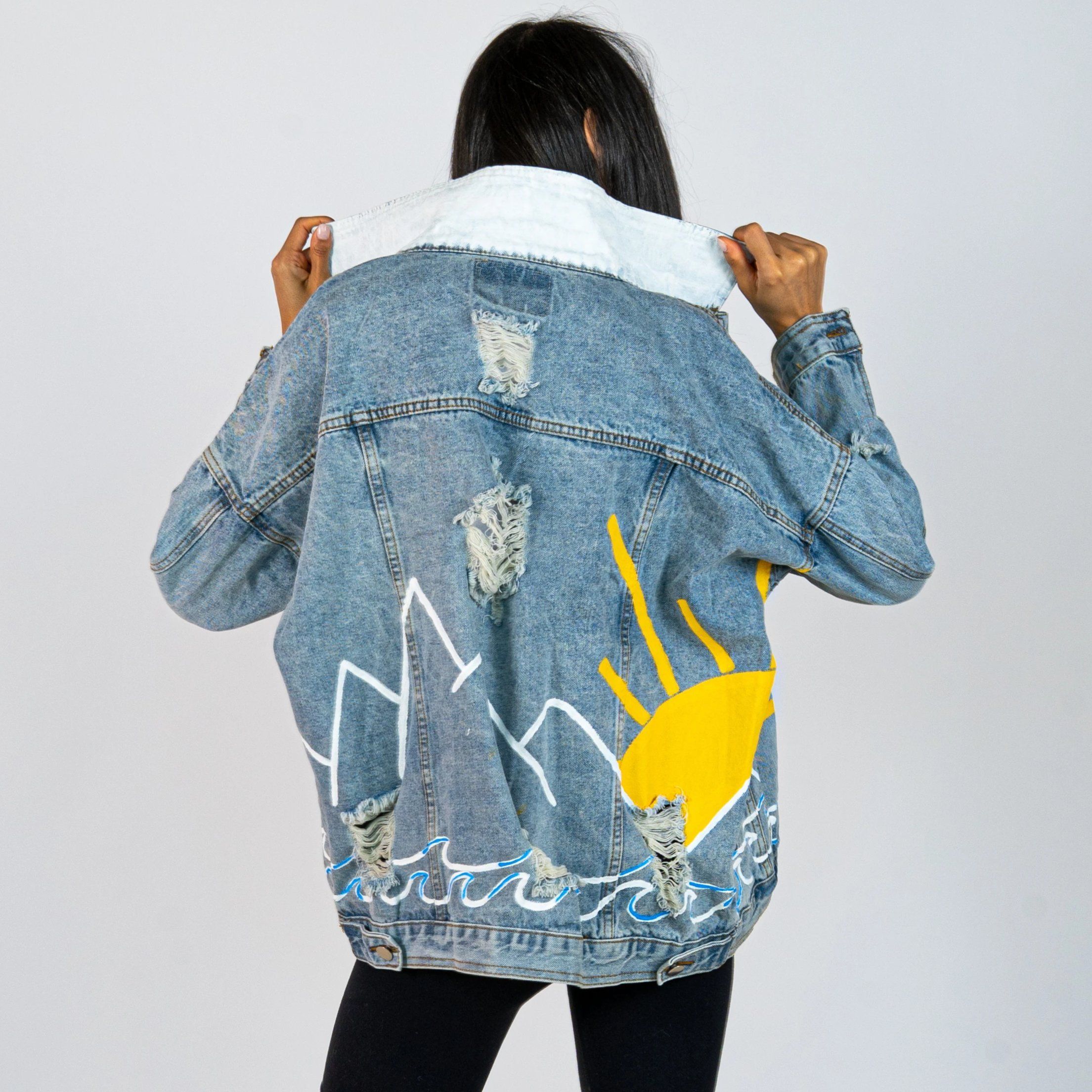 Lighter blue denim wash. Outline of mountain range, ocean waves, and sun setting surrounding entire jacket. Collar and front pockets painted white. Signed @wrenandglory.