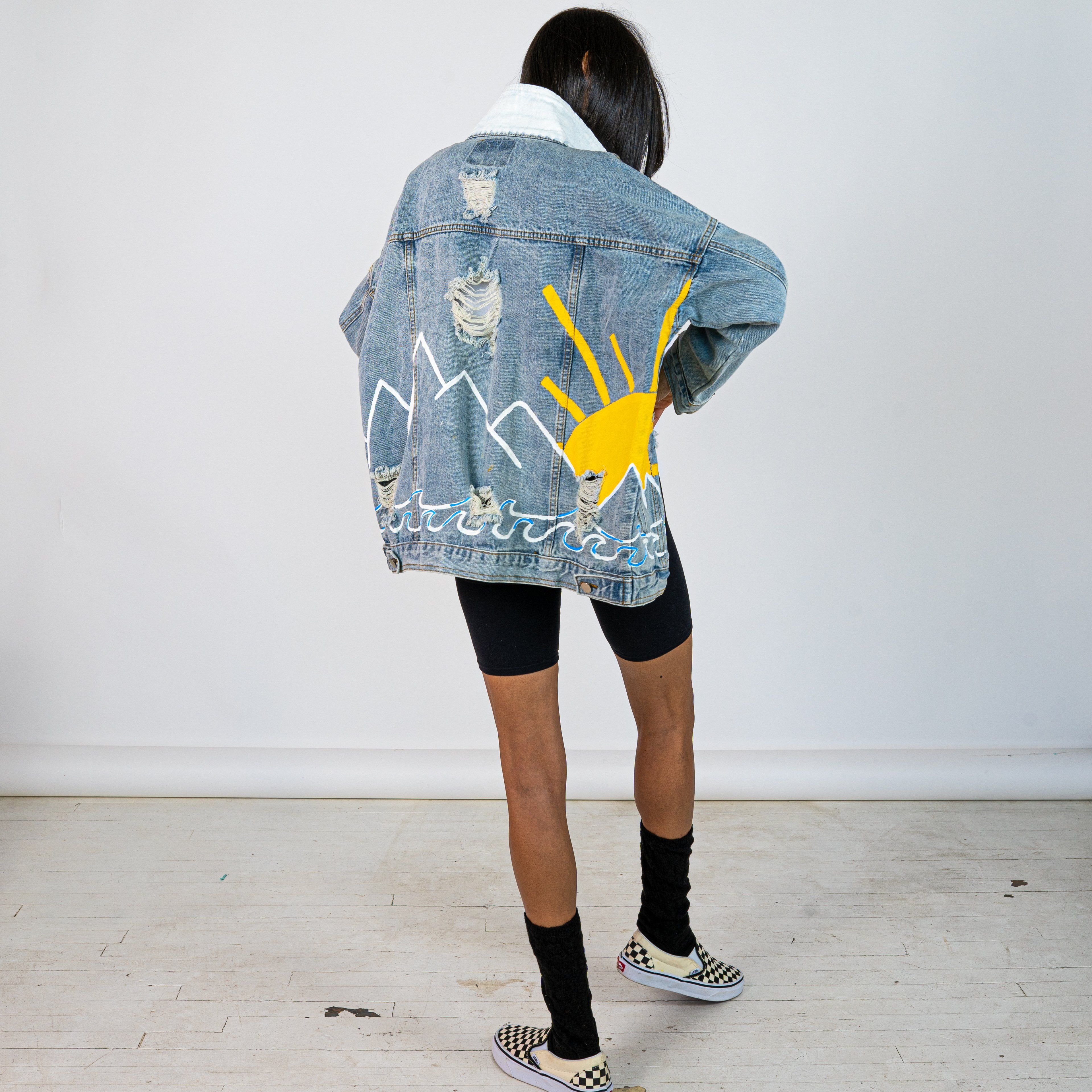Lighter blue denim wash. Outline of mountain range, ocean waves, and sun setting surrounding entire jacket. Collar and front pockets painted white. Signed @wrenandglory.