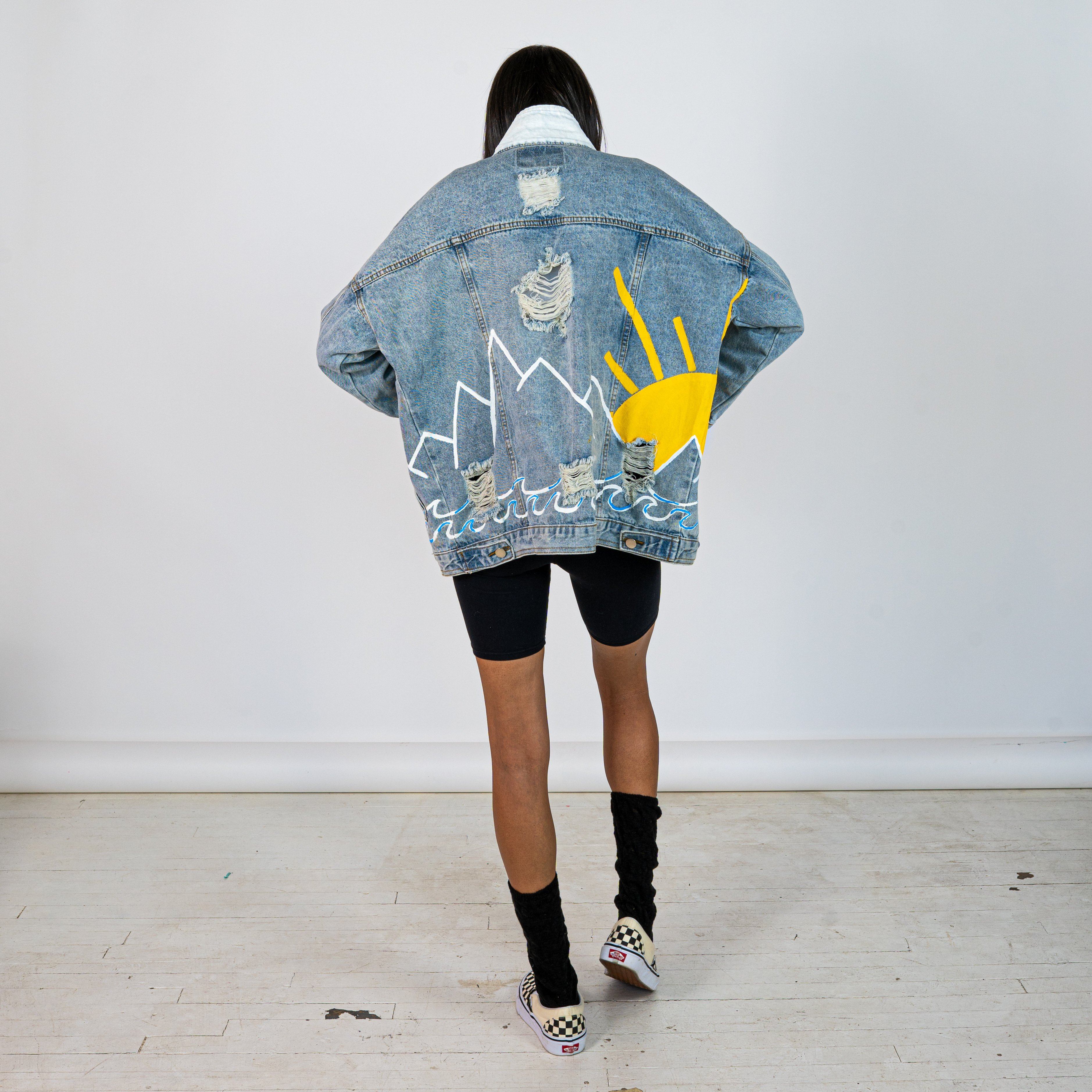 Lighter blue denim wash. Outline of mountain range, ocean waves, and sun setting surrounding entire jacket. Collar and front pockets painted white. Signed @wrenandglory.