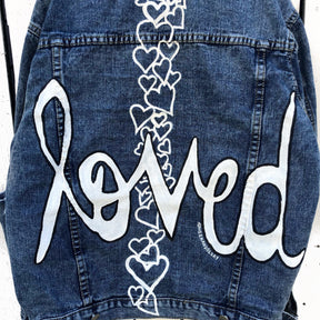Medium blue denim wash. 'Loved' painted in white, block cursive, outlines with black on lower back, with row of assorted sized heart clusters down the back center. Signed @wrenandglory.