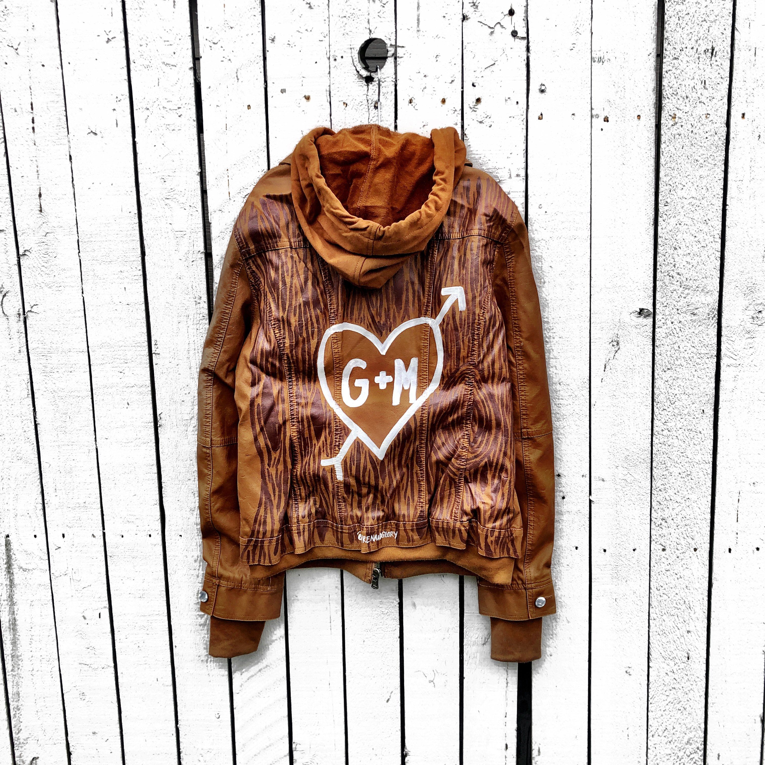 Light brown, faux leather jacket with tan sweatshirt lining the inside, with good and thumbholes at sleeves. Tree trunk painted across entire back in dark brown, with heart carved out and ' ___ + ___' painted inside. Please specify names above. Signed @wrenandglory.