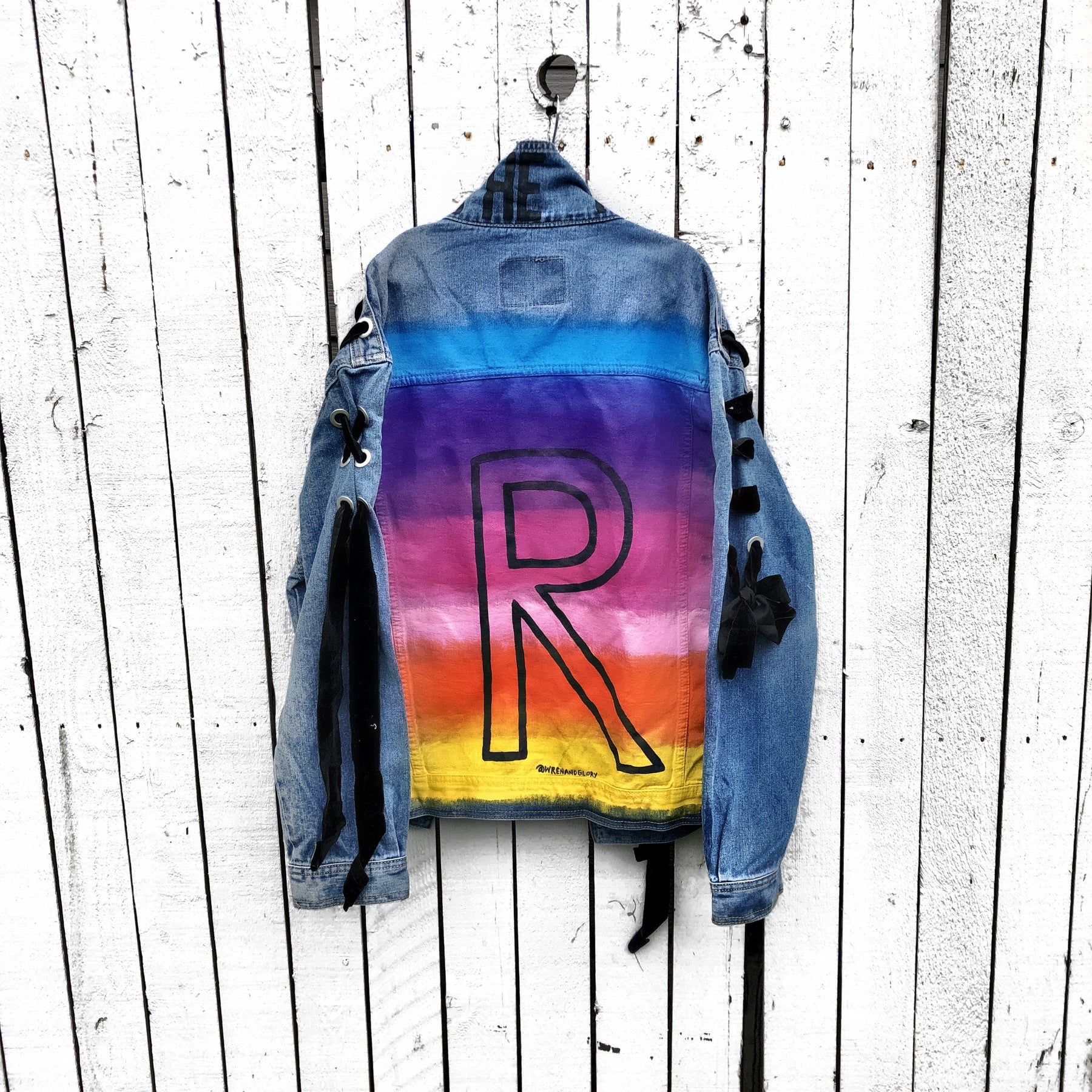 Dark blue denim wash, with velvert ribbon threaded through arms and front, lower left side. Ombre of colored striped on back, with large black initial painted in open block font over the colors. 'BE THE MAGIC' painted on color in black. Please specify initial above. Signed @wrenandglory.