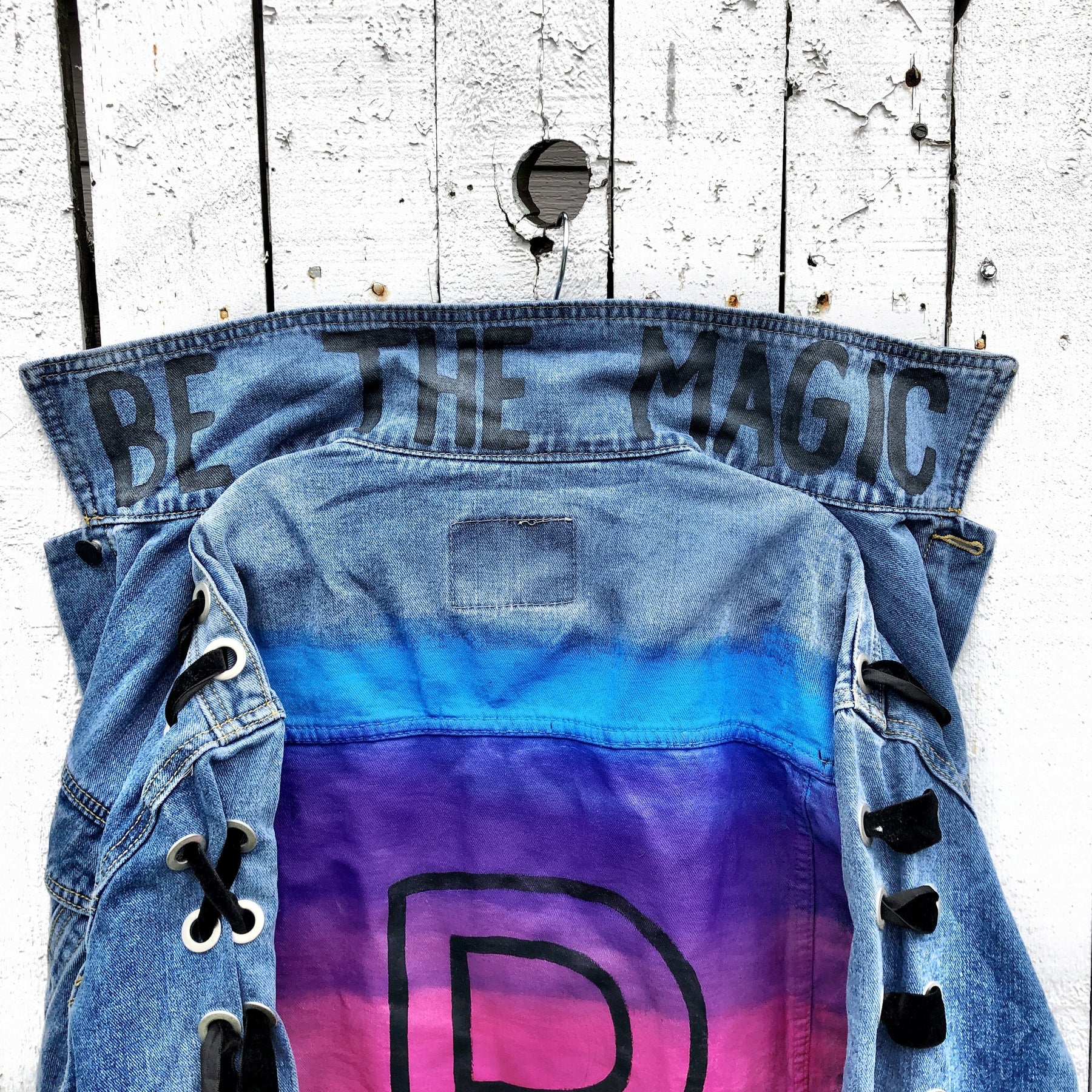Dark blue denim wash, with velvert ribbon threaded through arms and front, lower left side. Ombre of colored striped on back, with large black initial painted in open block font over the colors. 'BE THE MAGIC' painted on color in black. Please specify initial above. Signed @wrenandglory.