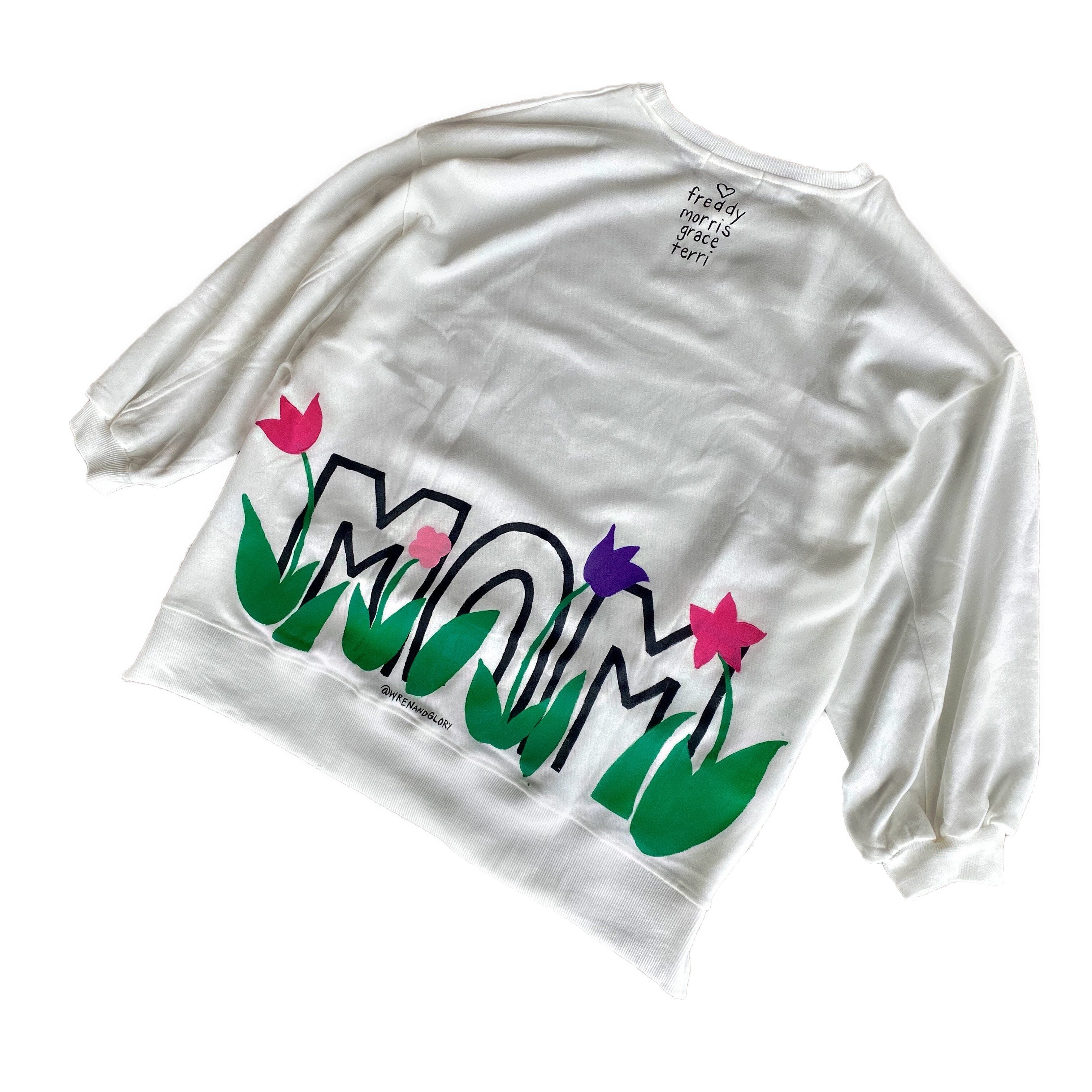 Oversized white sweatshirt. 'MOM' painted in on back, coming up from bottom hem, with flower pattern painted throughout letters. Customized upper back with names of children, grandchildren, husband, etc. Up to 8 names able to be painted. Small red embroidered heart on each lower sleeve. Signed @wrenandglory.