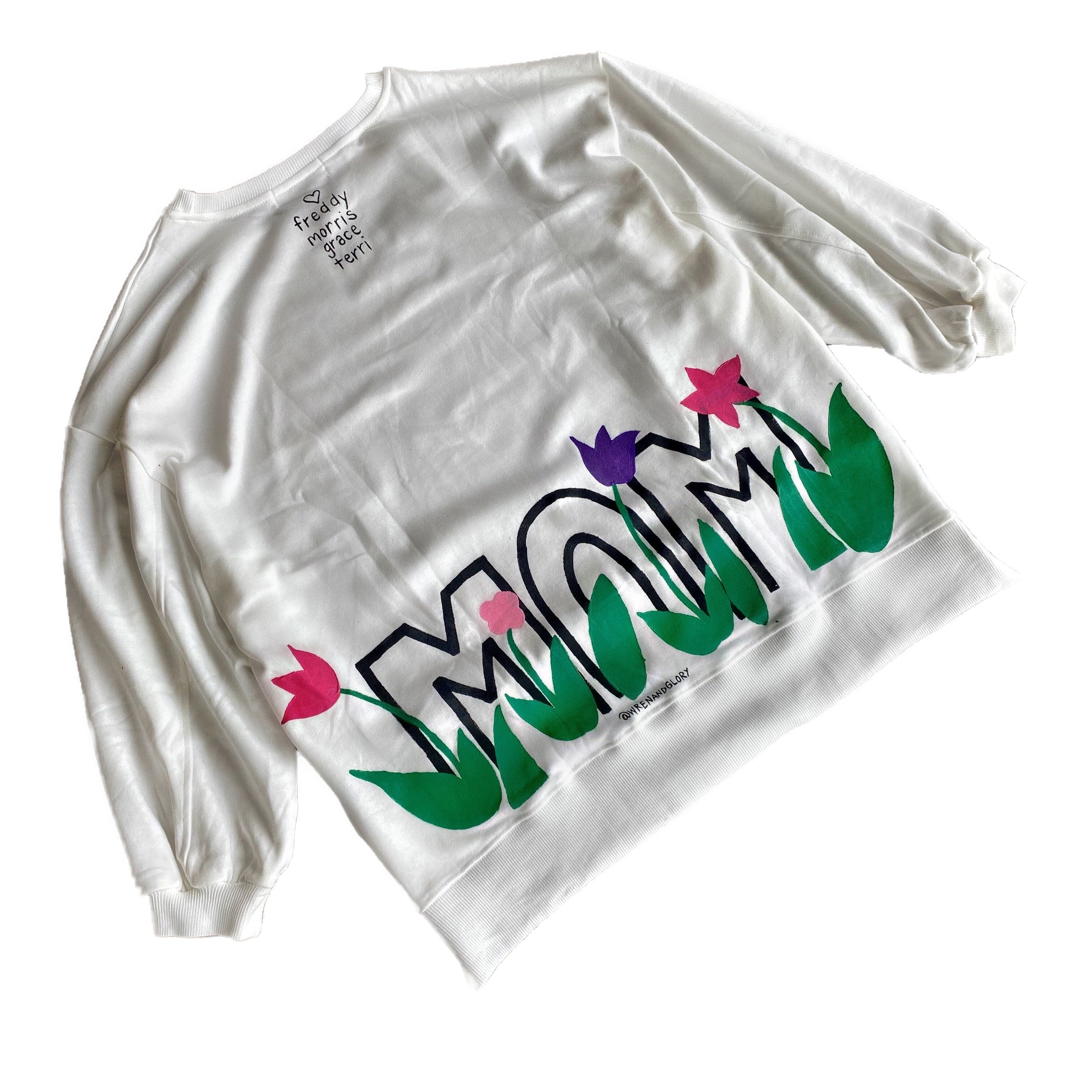 Oversized white sweatshirt. 'MOM' painted in on back, coming up from bottom hem, with flower pattern painted throughout letters. Customized upper back with names of children, grandchildren, husband, etc. Up to 8 names able to be painted. Small red embroidered heart on each lower sleeve. Signed @wrenandglory.