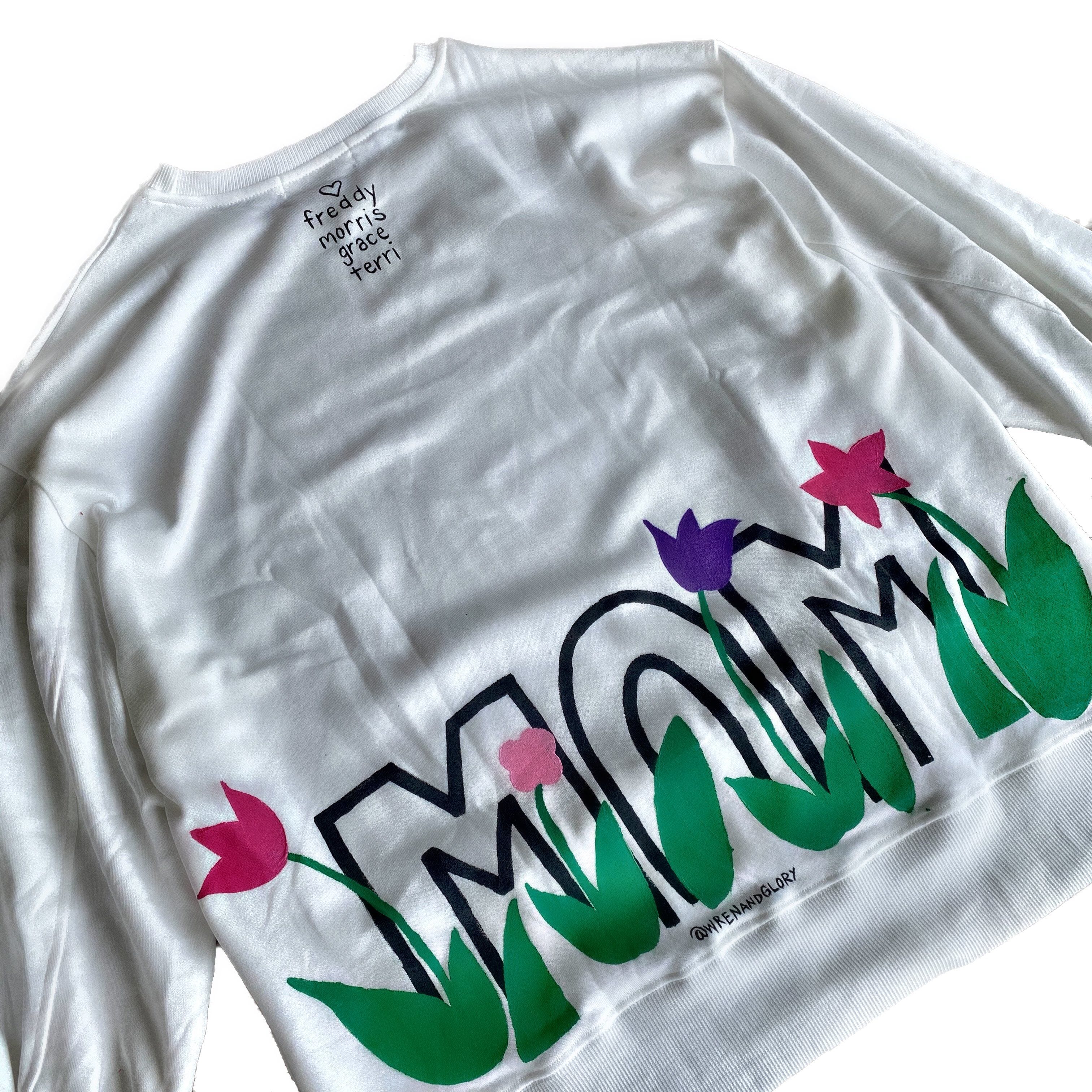 Oversized white sweatshirt. 'MOM' painted in on back, coming up from bottom hem, with flower pattern painted throughout letters. Customized upper back with names of children, grandchildren, husband, etc. Up to 8 names able to be painted. Small red embroidered heart on each lower sleeve. Signed @wrenandglory.