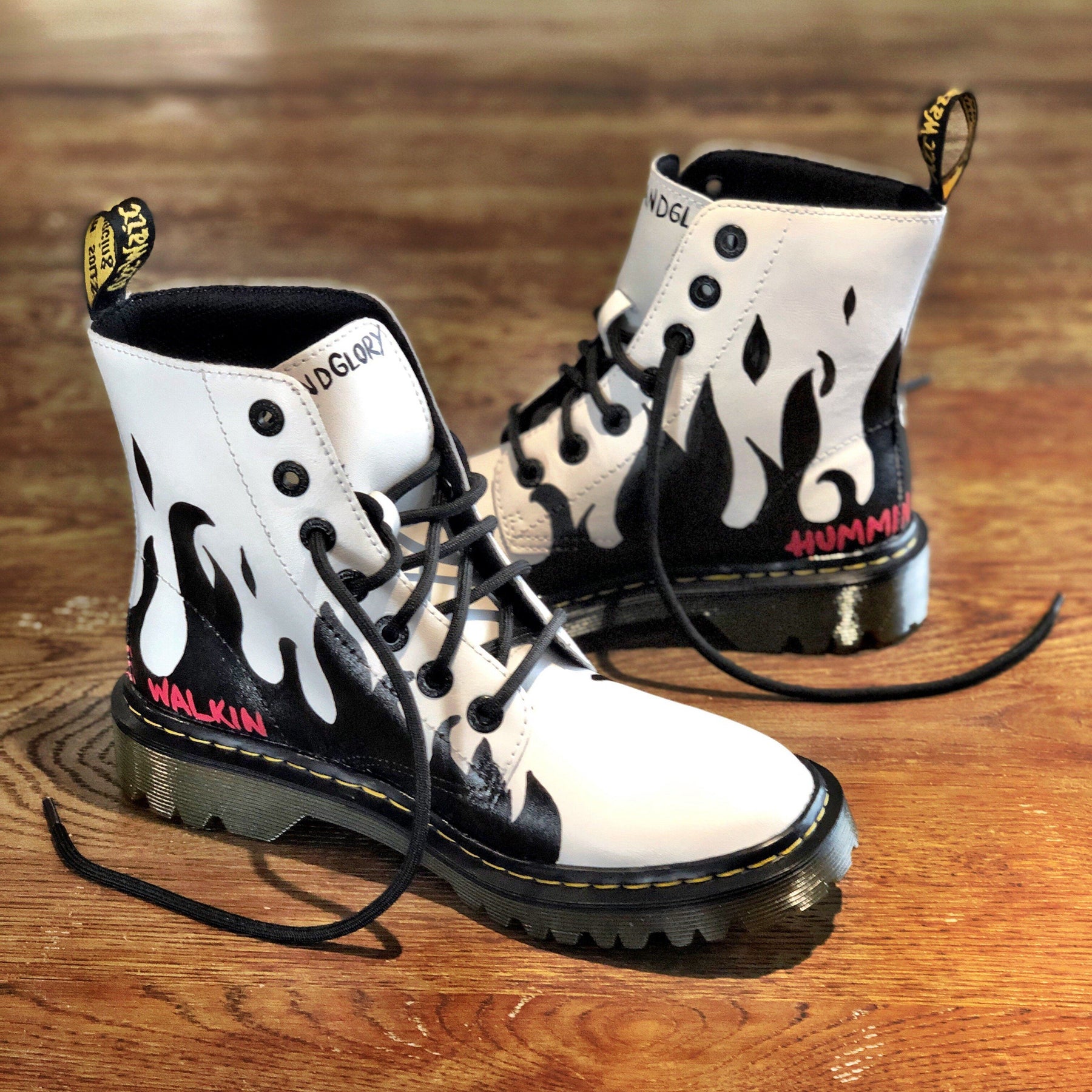 Black flames painted along the entire bottom of boots, with the words 'now you're walkin' painted in small font on one, and 'hummin your song' painted in small font on the other (in red on both). Painted on white Dr Marten boots.