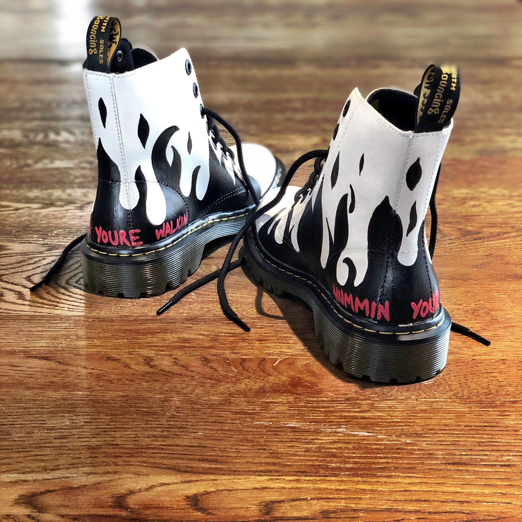 Black flames painted along the entire bottom of boots, with the words 'now you're walkin' painted in small font on one, and 'hummin your song' painted in small font on the other (in red on both). Painted on white Dr Marten boots.