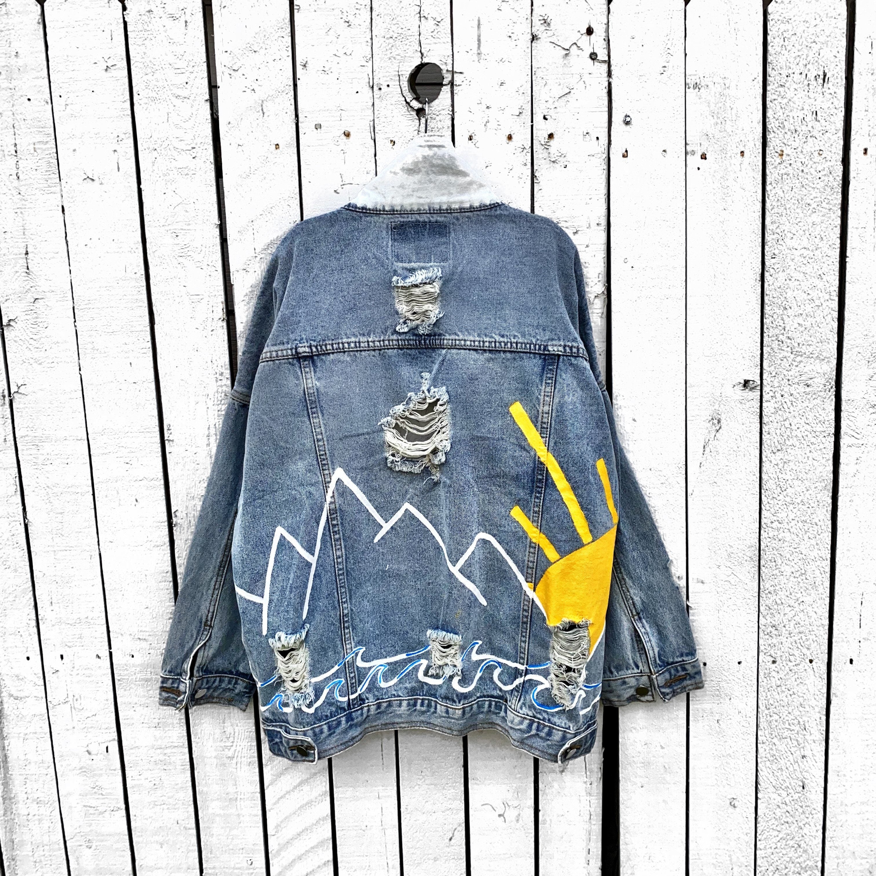 Lighter blue denim wash. Outline of mountain range, ocean waves, and sun setting surrounding entire jacket. Collar and front pockets painted white. Signed @wrenandglory.