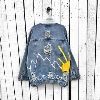 Lighter blue denim wash. Outline of mountain range, ocean waves, and sun setting surrounding entire jacket. Collar and front pockets painted white. Signed @wrenandglory.