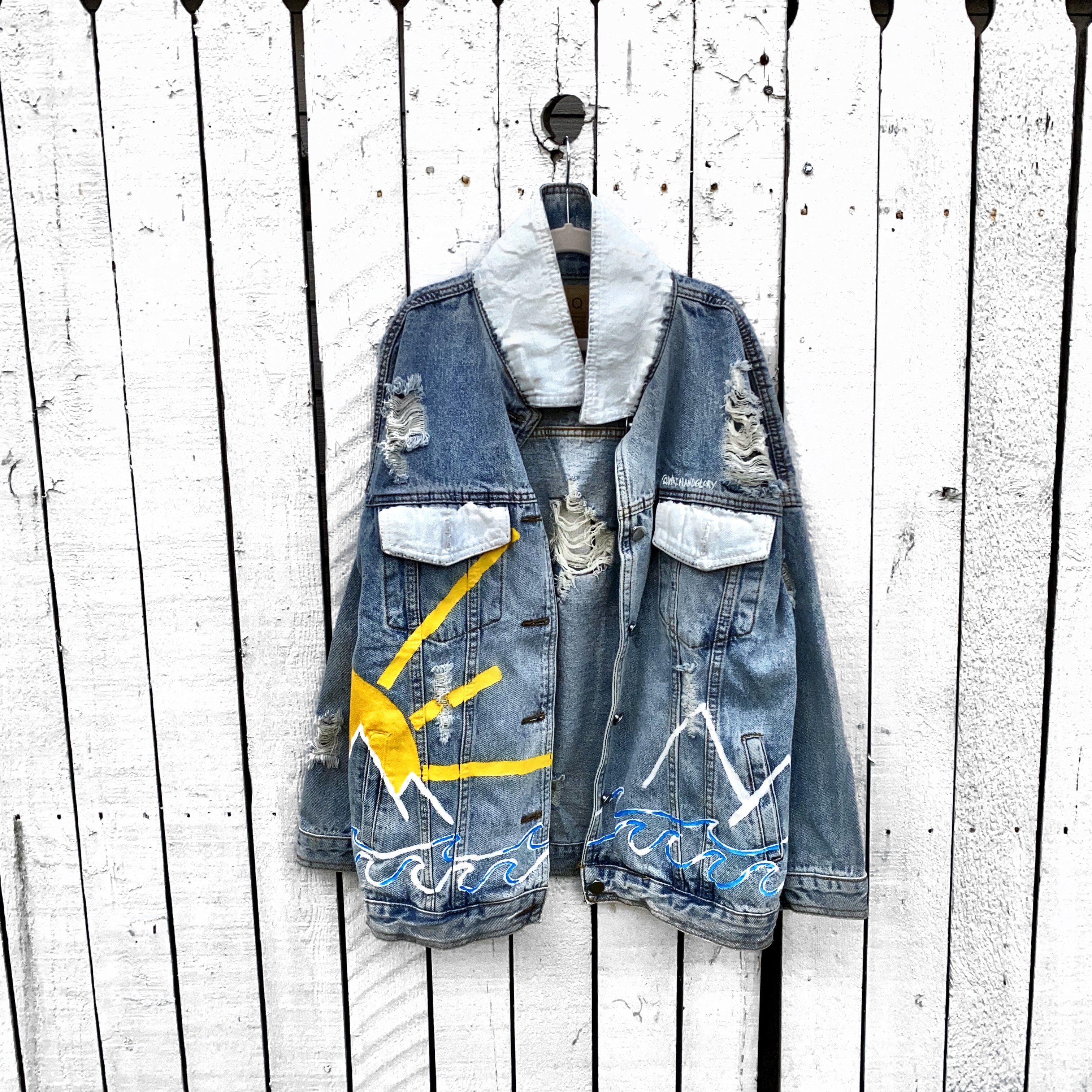 Lighter blue denim wash. Outline of mountain range, ocean waves, and sun setting surrounding entire jacket. Collar and front pockets painted white. Signed @wrenandglory.