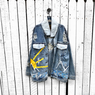 Lighter blue denim wash. Outline of mountain range, ocean waves, and sun setting surrounding entire jacket. Collar and front pockets painted white. Signed @wrenandglory.