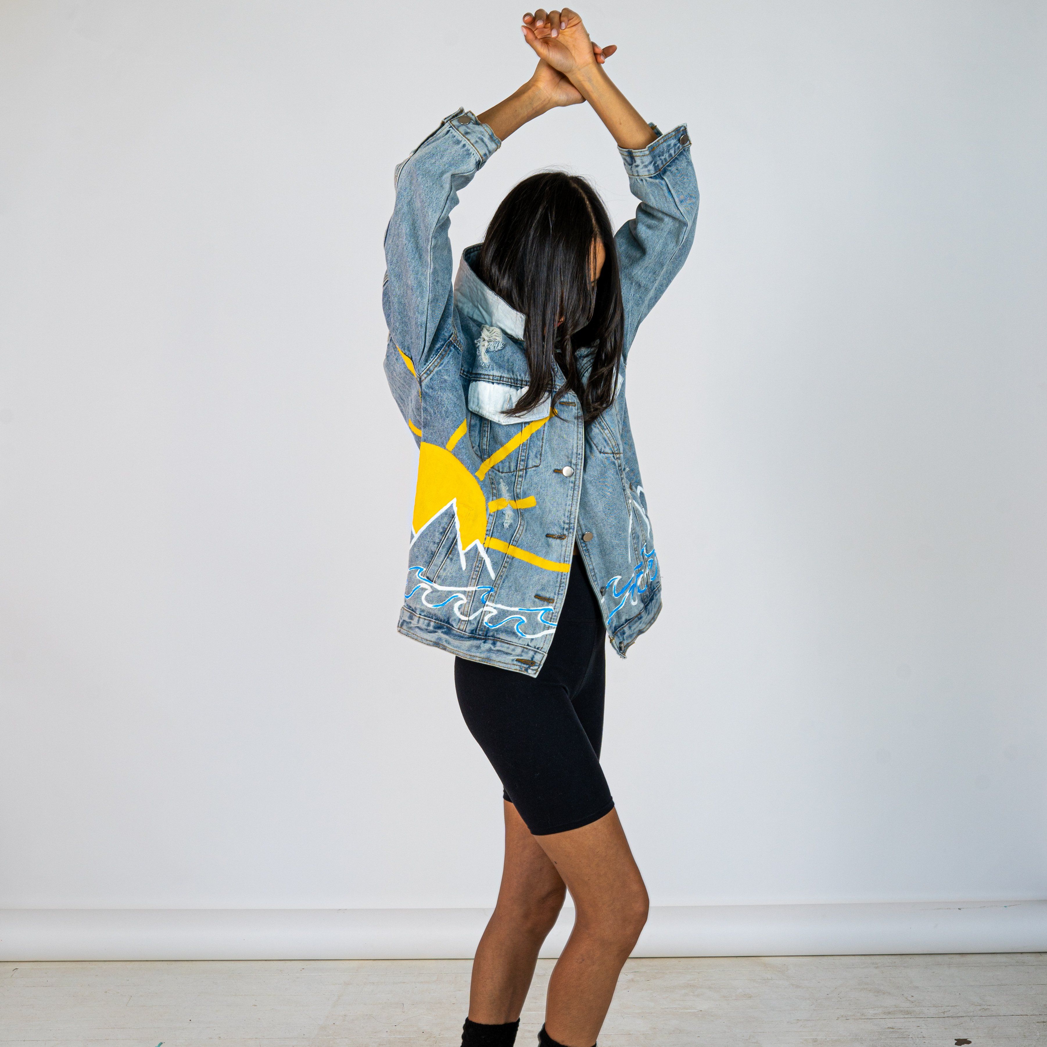 Lighter blue denim wash. Outline of mountain range, ocean waves, and sun setting surrounding entire jacket. Collar and front pockets painted white. Signed @wrenandglory.