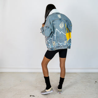 Lighter blue denim wash. Outline of mountain range, ocean waves, and sun setting surrounding entire jacket. Collar and front pockets painted white. Signed @wrenandglory.