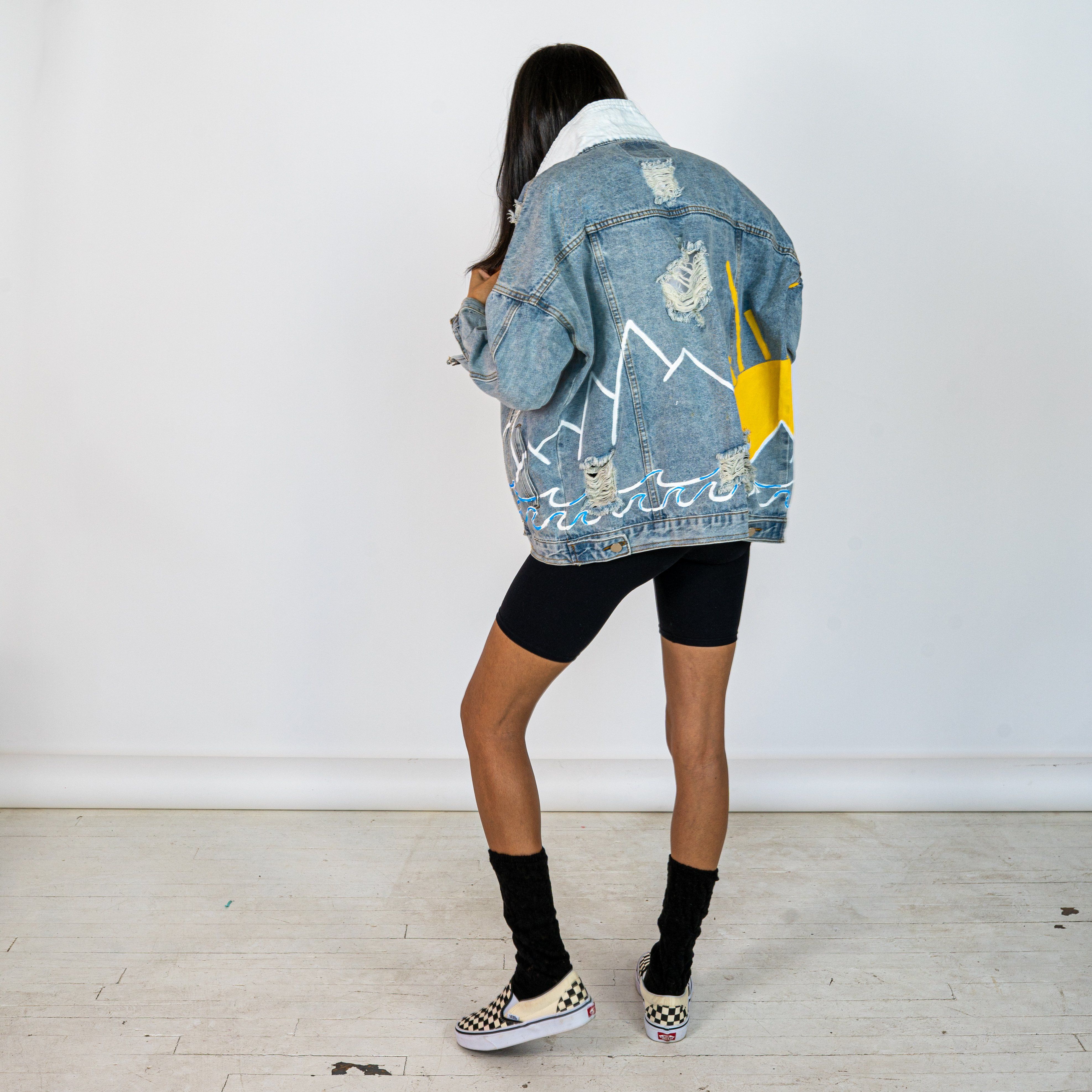 Lighter blue denim wash. Outline of mountain range, ocean waves, and sun setting surrounding entire jacket. Collar and front pockets painted white. Signed @wrenandglory.