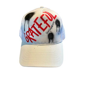 White trucker hat. GRATEFUL painted on hat in red, with black spray paint. Signed @wrenandglory.