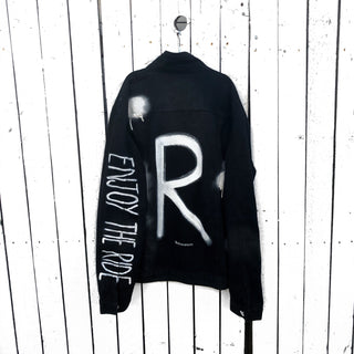 Black denim. Initial on back in white, with white and black painted 'splatters' 'Enjoy The Ride' painted in white down the arm Signed @wrenandglory.