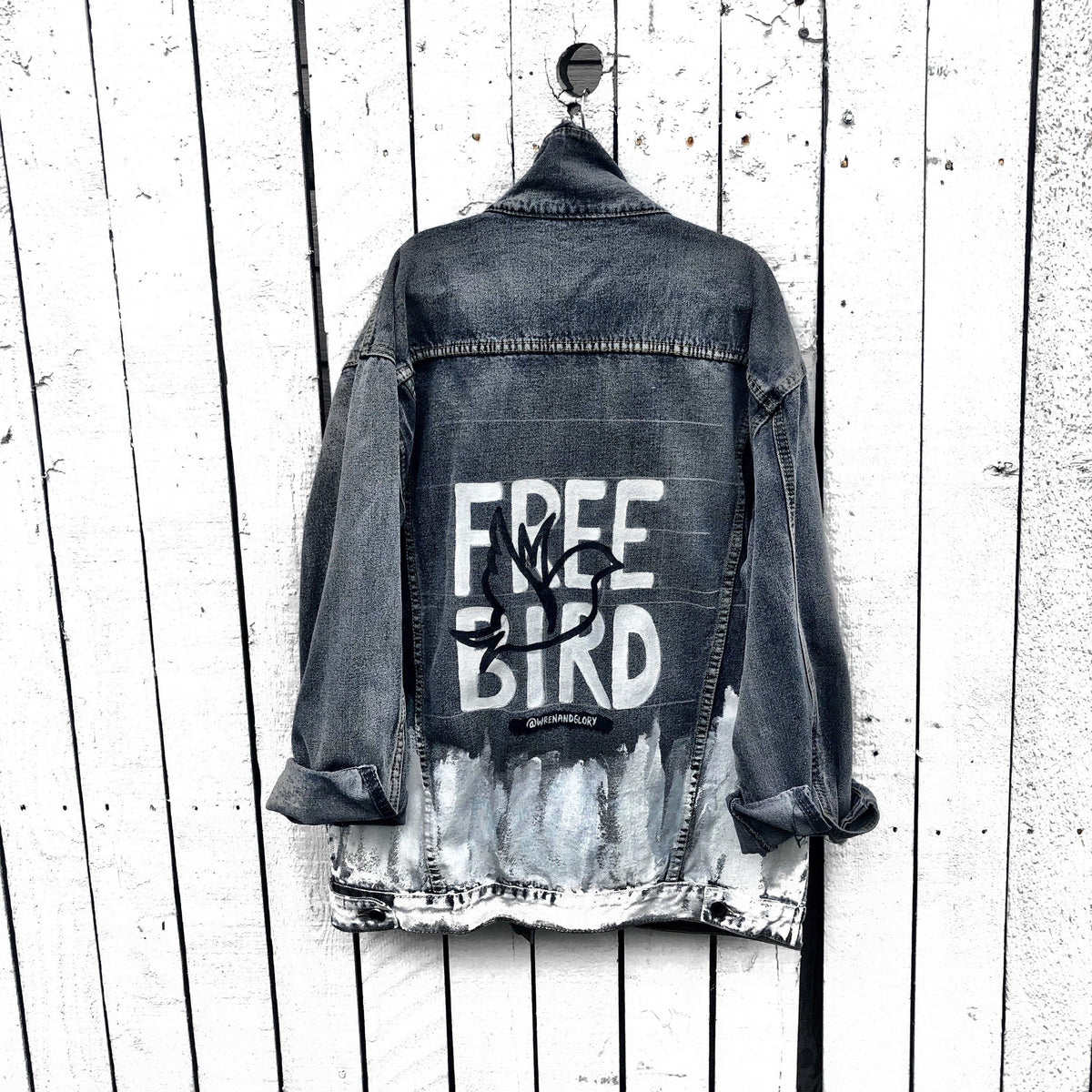 Faded black denim. FREE BIRD painted in white in block letters on back, with a silhouette on a bird in black over it. Signed @wrenandglory.