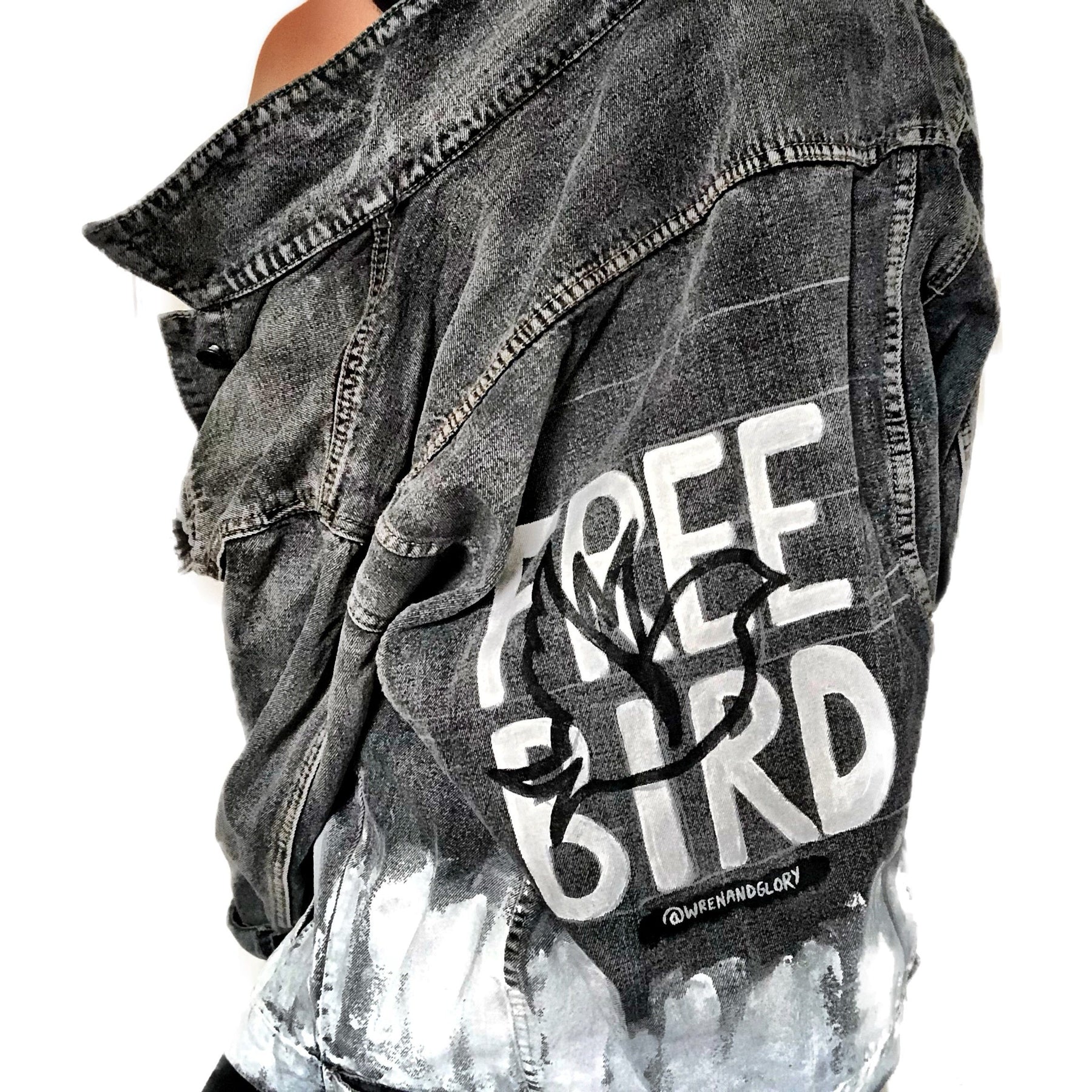 Faded black denim. FREE BIRD painted in white in block letters on back, with a silhouette on a bird in black over it. Signed @wrenandglory.