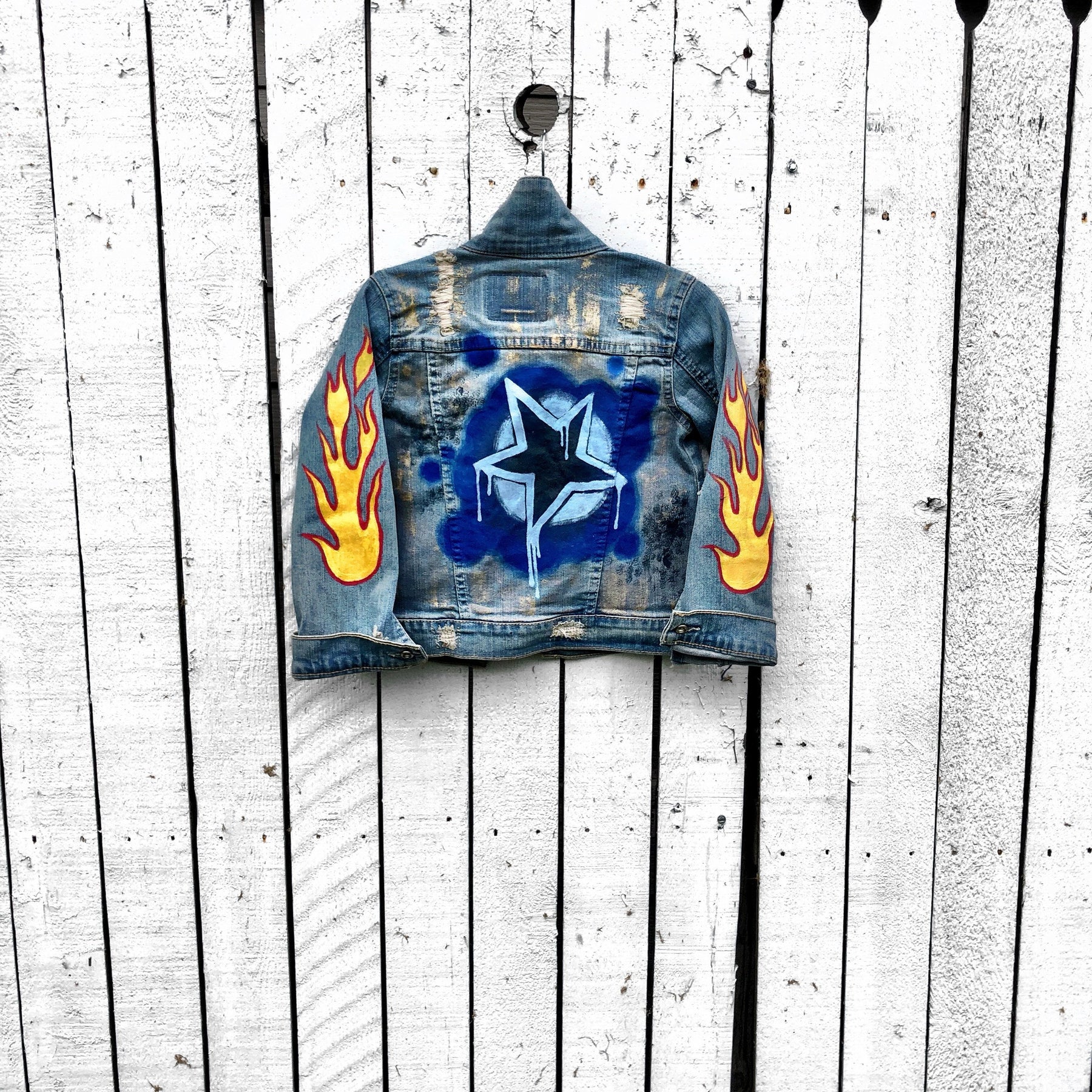 Blue denim jacket. Hand painted design, with stars, flames, and metallics.  Childs name available on front, left corner. Each jacket wrapped in a silk wrap bracelet with a child’s charm. Each piece signed @wrenandglory.