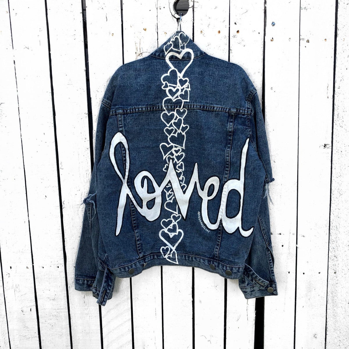 Medium blue denim wash. 'Loved' painted in white, block cursive, outlines with black on lower back, with row of assorted sized heart clusters down the back center. Signed @wrenandglory.