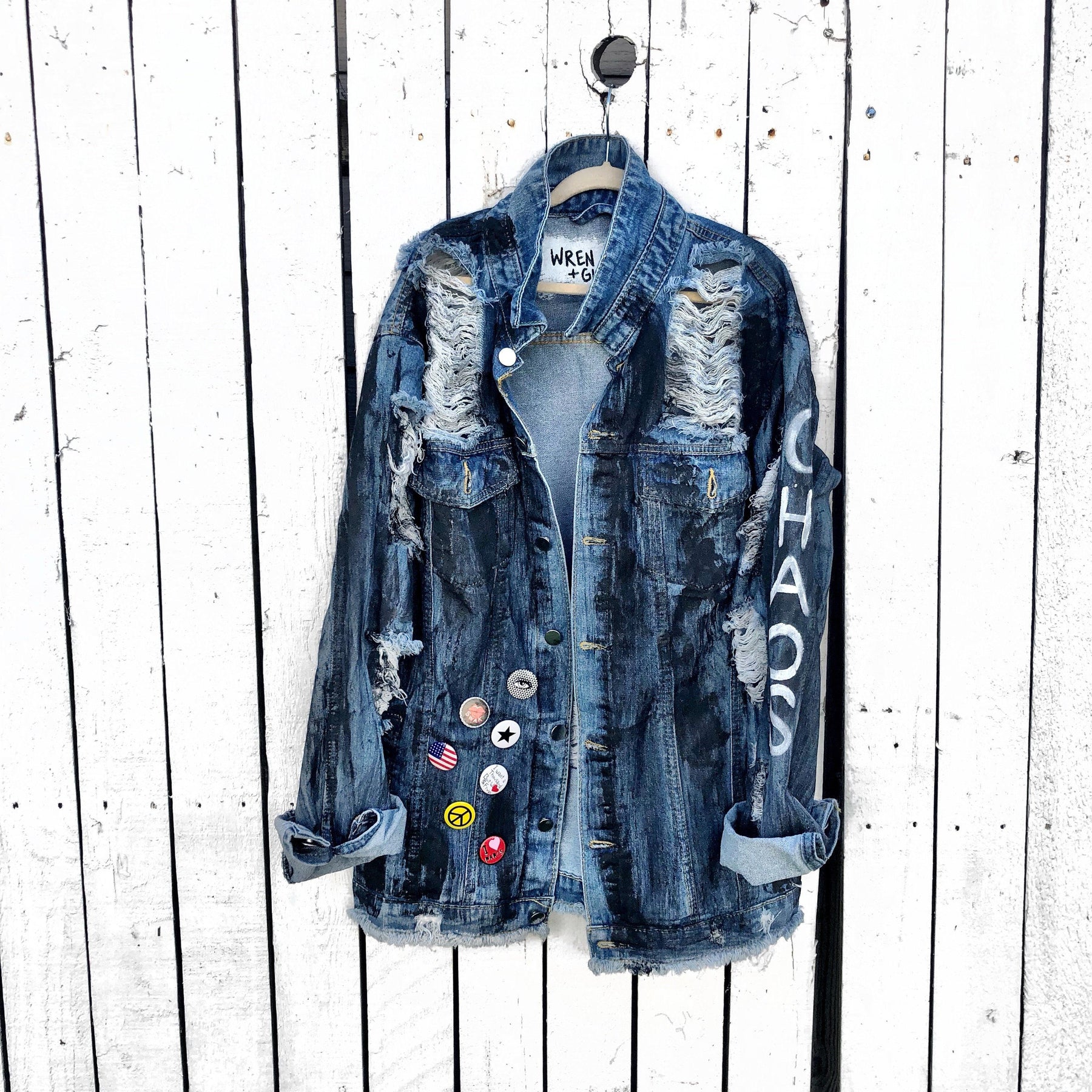 Medium blue denim wash. Black base all over, with 'MADE IN NYC' painted on back in white. CHAOS painted down the arm in white. Assorted 1" pins on bottom left, bottom pocket area. Signed @wrenandglory.