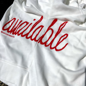 Oversized white sweatshirt. 'Available' painted in script on lower left side, in red. Small red embroidered heart on each lower sleeve. Signed @wrenandglory.