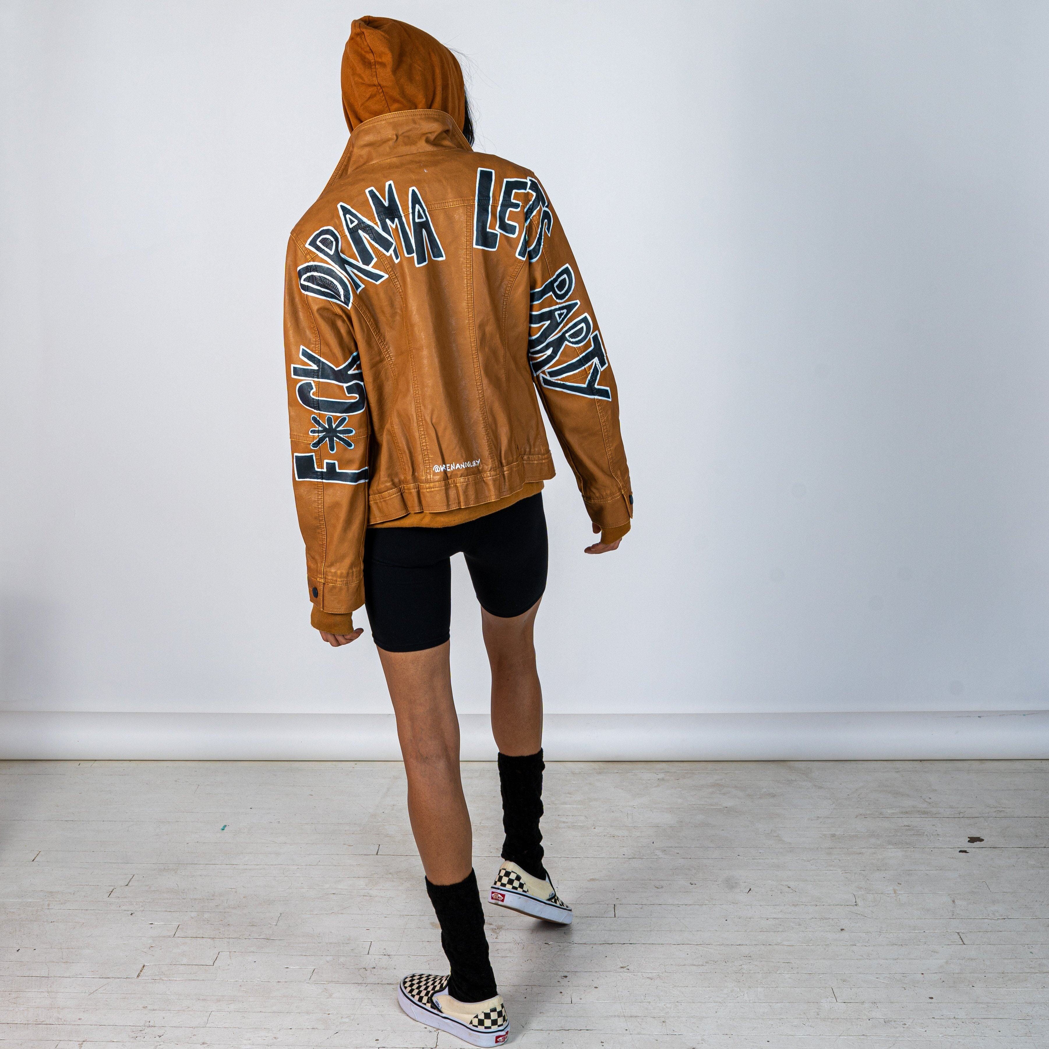 Tan, faux leather jacket with Brown hooded sweatshirt lining the inside and thumbholes at sleeves. 'F*CK DRAMA LET'S PARTY' painted along back of sleeves and upper back of jacket, in black, with white outline. Signed @wrenandglory.