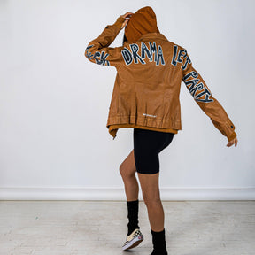 Tan, faux leather jacket with Brown hooded sweatshirt lining the inside and thumbholes at sleeves. 'F*CK DRAMA LET'S PARTY' painted along back of sleeves and upper back of jacket, in black, with white outline. Signed @wrenandglory.