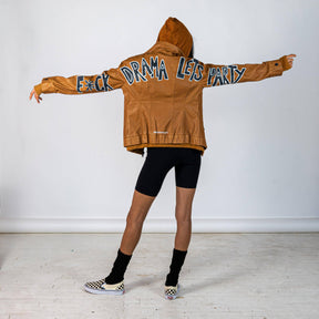 Tan, faux leather jacket with Brown hooded sweatshirt lining the inside and thumbholes at sleeves. 'F*CK DRAMA LET'S PARTY' painted along back of sleeves and upper back of jacket, in black, with white outline. Signed @wrenandglory.