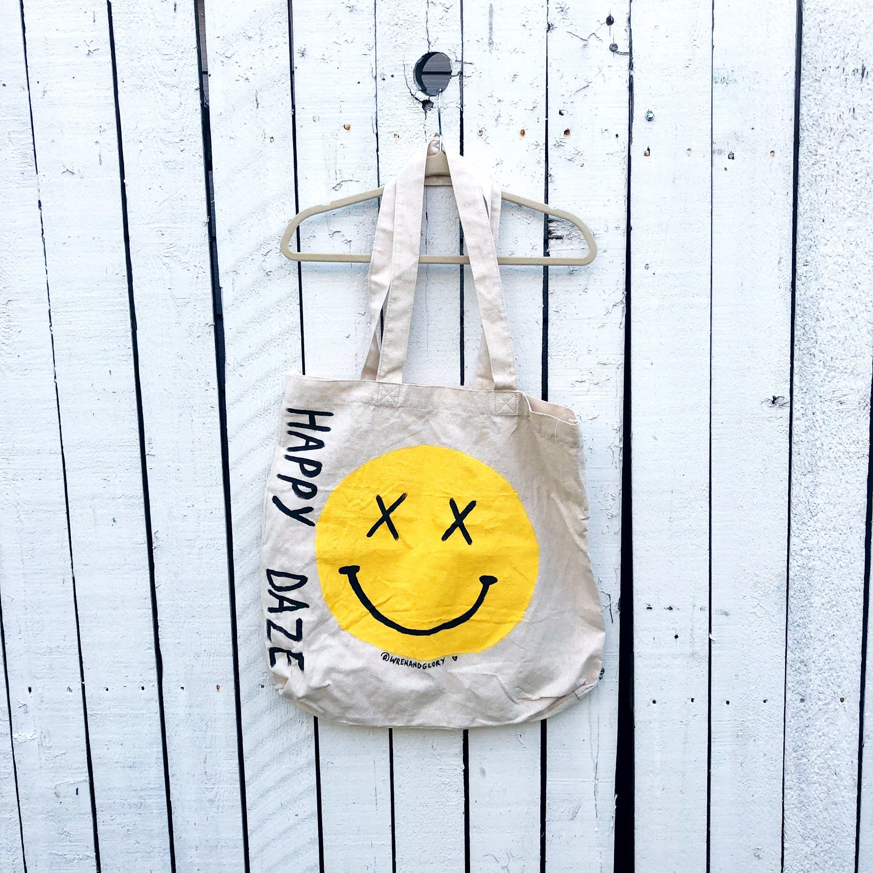 Off white canvas tote bag. Hand painted 'HAPPY DAZE' along the side, with a large yellow smiley in center.