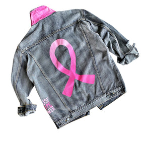 Faded black denim. Pink ombre Breast Cancer ribbon painted in large on back. Love, Strength, Trust, Beauty, Peace, Hope, Passion, Faith, Power painted in light & bright pink on the front. Collar painted in pink ombre. Signed @wrenandglory.