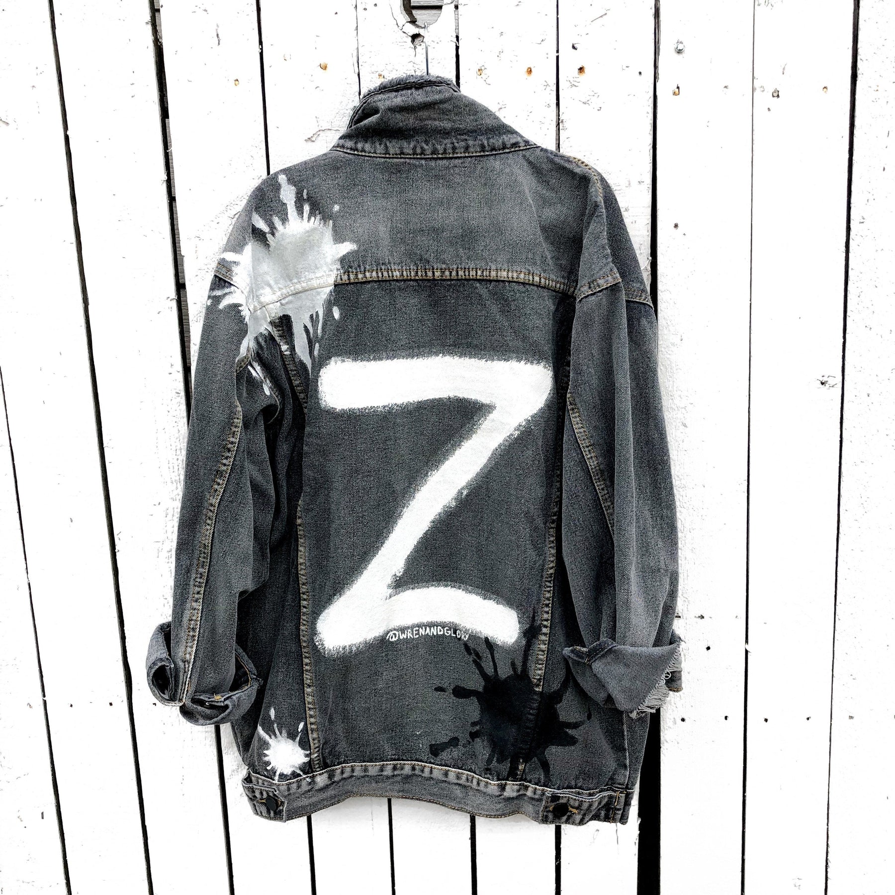 Faded black denim wash. Large white initial on back, with painted white and black 'splatters' 'Enjoy The Ride' painted in black and white down the arm. Signed @wrenandglory.