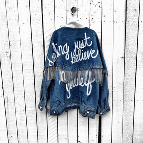 Darker blue denim wash with silver chains. 'DARLING JUST BELIEVE IN YOURSELF' painted in white script on back. Color and front pockets painted white. Signed @wrenandglory.