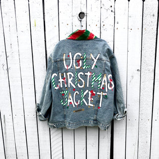 Lighter blue denim wash. UGLY CHRISTMAS JACKET painted in white, with black outline, on back. Ornaments, candy canes, mistletoe and other holiday related items decorating the letters. Collar and front pockets painted red and green, with gold boarders. Signed @wrenandglory.