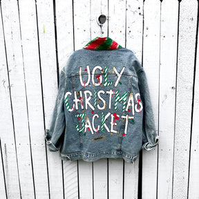 Lighter blue denim wash. UGLY CHRISTMAS JACKET painted in white, with black outline, on back. Ornaments, candy canes, mistletoe and other holiday related items decorating the letters. Collar and front pockets painted red and green, with gold boarders. Signed @wrenandglory.