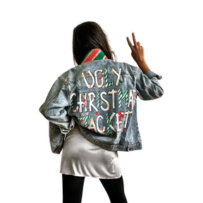 Lighter blue denim wash. UGLY CHRISTMAS JACKET painted in white, with black outline, on back. Ornaments, candy canes, mistletoe and other holiday related items decorating the letters. Collar and front pockets painted red and green, with gold boarders. Signed @wrenandglory.