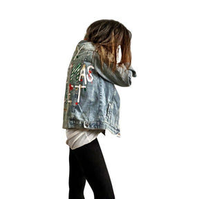 Lighter blue denim wash. UGLY CHRISTMAS JACKET painted in white, with black outline, on back. Ornaments, candy canes, mistletoe and other holiday related items decorating the letters. Collar and front pockets painted red and green, with gold boarders. Signed @wrenandglory.