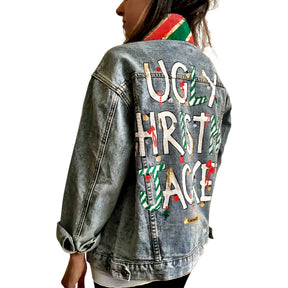 Lighter blue denim wash. UGLY CHRISTMAS JACKET painted in white, with black outline, on back. Ornaments, candy canes, mistletoe and other holiday related items decorating the letters. Collar and front pockets painted red and green, with gold boarders. Signed @wrenandglory.