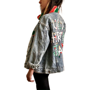 Lighter blue denim wash. UGLY CHRISTMAS JACKET painted in white, with black outline, on back. Ornaments, candy canes, mistletoe and other holiday related items decorating the letters. Collar and front pockets painted red and green, with gold boarders. Signed @wrenandglory.