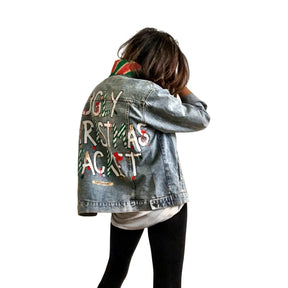Lighter blue denim wash. UGLY CHRISTMAS JACKET painted in white, with black outline, on back. Ornaments, candy canes, mistletoe and other holiday related items decorating the letters. Collar and front pockets painted red and green, with gold boarders. Signed @wrenandglory.