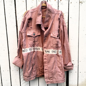 Light blush colored denim. Large eye painted on back, with lines following the shape. A banner spanning the entire jacket painted in white, with black wording. Signed @wrenandglory.
