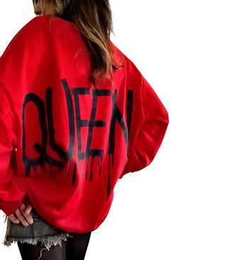 A person wearing an oversized red sweatshirt with 'QUEEN' painted in black on the back and a black crown painted on the front, upper left side.