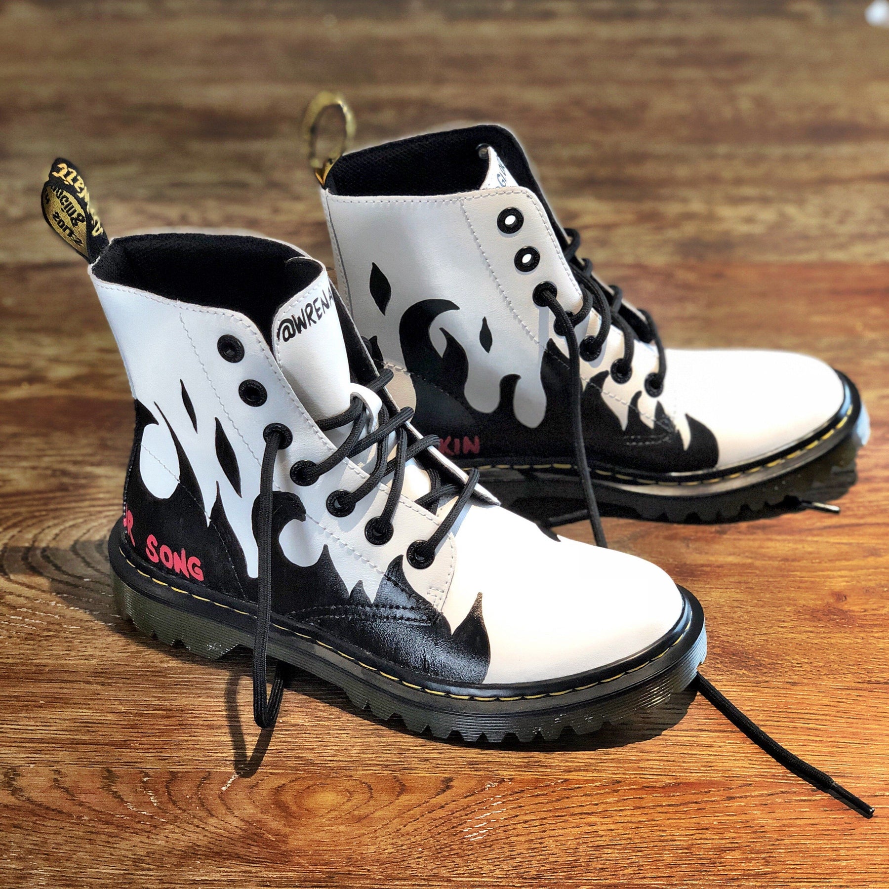 Black flames painted along the entire bottom of boots, with the words 'now you're walkin' painted in small font on one, and 'hummin your song' painted in small font on the other (in red on both). Painted on white Dr Marten boots.